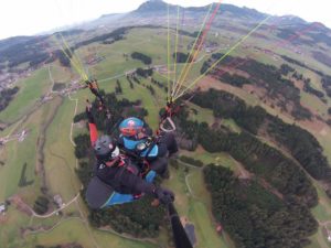 Das Bild zeigt einen Tandemflug mit dem Gleitschirm. dabei hängt der Passagier von dem Piloten. Das Foto wurde von oben aufgenommen